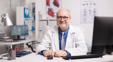 Obraz premium Mature doctor wearing eyeglasses and smiling in hospital cabinet sitting at desk.