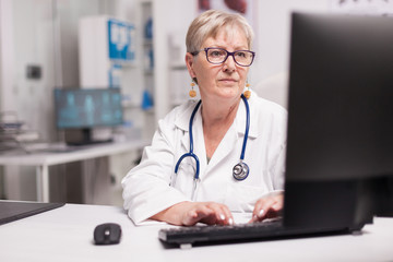 Mature doctor typing patient report on computer in hospital cabinet wearing stethoscope around the...