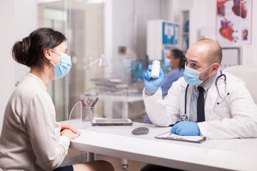 Doctor wearing protection mask against covid 19 showing bottle of pills to patient. Medic wearing white coat and stethoscope.