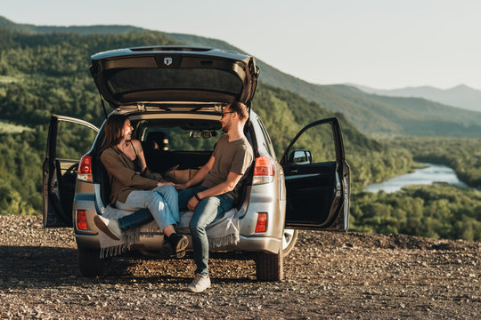 Young Traveler Couple on a Road Trip, Man and Woman Sitting on the Opened Trunk of Their Car Over Sunset