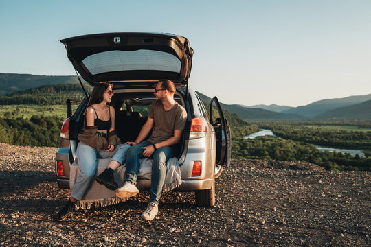 Young Traveler Couple on a Road Trip, Man and Woman Sitting on the Opened Trunk of Their Car Over Sunset