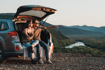 Young Traveler Couple on a Road Trip, Man and Woman Sitting on the Opened Trunk of Their Car Over...