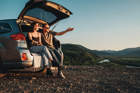 Young Traveler Couple on a Road Trip, Man and Woman Sitting on the Opened Trunk of Their Car Over Sunset