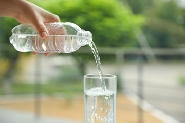 Woman pouring water from bottle into glass against blurred background, closeup