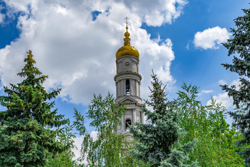 Fototapeta na wymiar Bell tower with the clock of the Holy Dormition Cathedral in Kharkiv (Ukraine) close-up. Tower of an Orthodox church with a golden dome and a cross against a background of blue sky and green trees