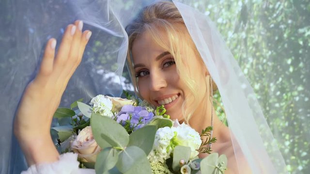 bride under veil with bouquet of flowers walks in Park.