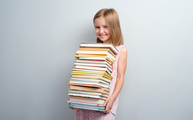 Smiling school girl holding stack of books on a grey wall background