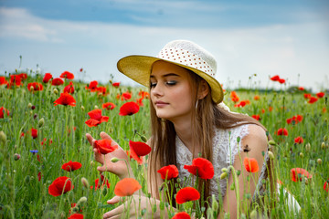 Portrait of a beautiful teenage girl in a field with wild poppies in a hat admiring the flowers