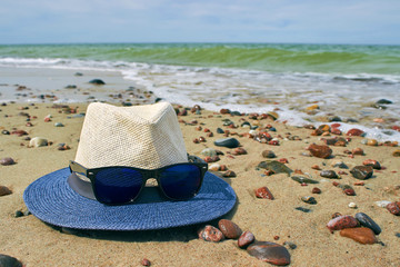 Straw hat and sunglasses lie on a sandy seashore with pebbles