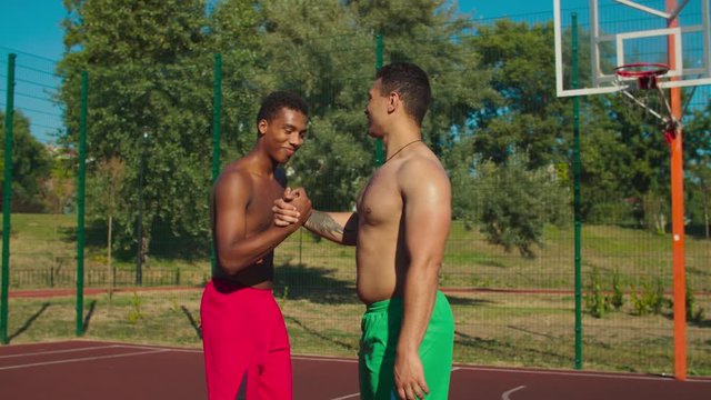 Cheerful shirtless athletic african american streetball players exchanging handshake on outdoor court after basketball game, expressing positivity, friendship, mutual respect and good sportsmanship.