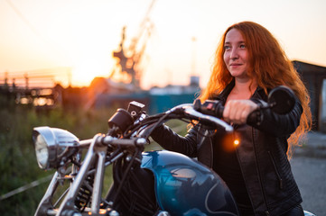 Fototapeta na wymiar Curly red-haired woman in a black leather jacket sits on a motorcycle at sunset. Portrait of a serious girl driving a bike.