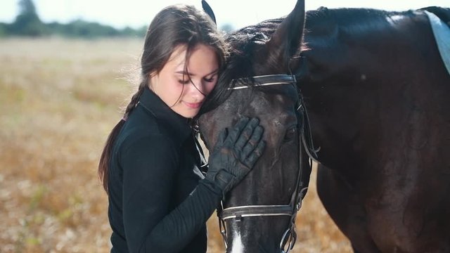 Young female jockey embracing her horse in the field at sunny daytime.