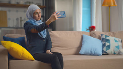 Obraz premium Arab Girl In Hijab Taking Photo On Smartphone Relaxing On Comfortable Couch In Living Room
