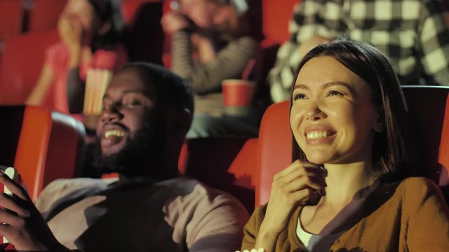 Close-up portrait of beautiful asian woman and handsome bearded dark-skinned man eating popcorn and laughing during funny movie. Multi-ethnic viewers having fun while watching comedy in cinema theater