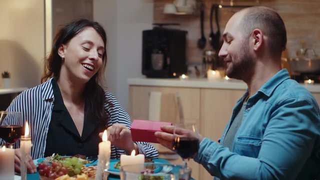 Surprised young woman with charming smile sitting at the table in kitchen opening small gift box present. Cheerful couple dining together at home, enjoying the meal, celebrating their anniversary