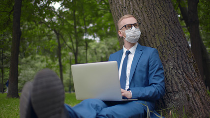 Young businessman working on laptop while sitting on grass under tree wearing protective mask