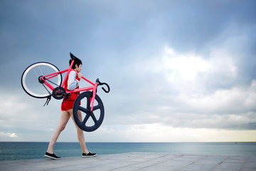Young sporty woman dressed in flamboyant clothing hold her light weight fixed gear bicycle on the shoulders while standing at seashore against cloudy sky copy space area for your text message content