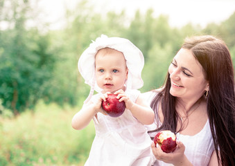 Happy young mother with a baby and red apple outdoor in summer day in a  white dresses