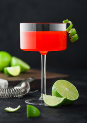 Cosmopolitan cocktail in modern crystal glass with lime peel and fresh limes with strainer on black background.