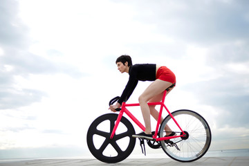 Fototapeta premium Young female cyclist riding on fixed gear bike against grey sky background with copy space area for your text message or advertising content, sporty woman enjoy ride on bicycle in cloudy day outdoors
