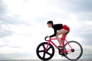 Full length portrait of young woman cyclist dressed in sexy red shorts riding bicycle along seashore against gray cloudy sky background with copy space area for your text message or advertise content