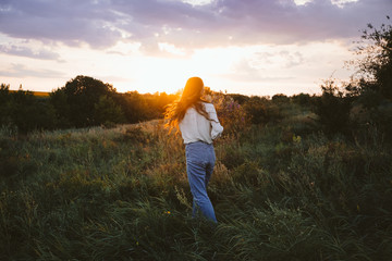 National Relaxation Day, relaxation practices, mental health, slow living concept. Young girl with long windy hair and flower bouquet enjoying nature on the background of sunset