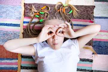 A Caucasian child, a cheerful girl with pigtails, lies on the floor on a woven striped rug, smiles,...