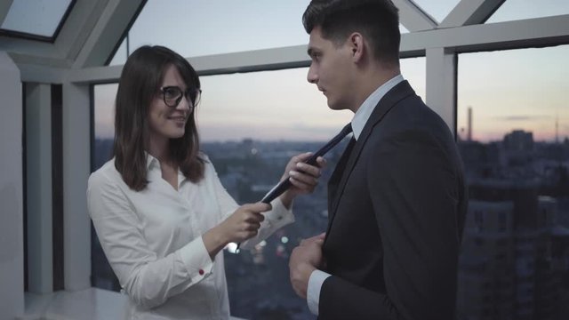 Portrait of young beautiful Caucasian woman flirting with handsome elegant man in office in the evening. Smiling brunette lady touching necktie of shy guy in office center in dusk. Work romance.