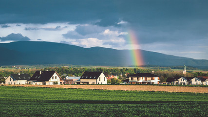 A stormy front over a mountain range, a colorful rainbow over the houses after the storm.