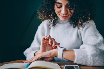 Positive curly female journalist checking time at electronic smartwatch waiting colleague for...