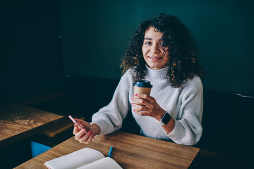Portrait of smiling hipster girl sitting at coffee shop holding mobile phone and waiting for call from friend, young cheerful woman enjoying caffeine beverage communicated via smartphone at campus
