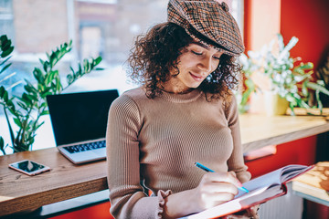 Young trendy woman writing organisation plan in textbook after e learning on netbook spending time at coworking space, positive female journalist creating list of meetings and interviews for next week