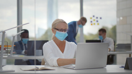 Mature businesswoman working on computer in protective mask sitting in office