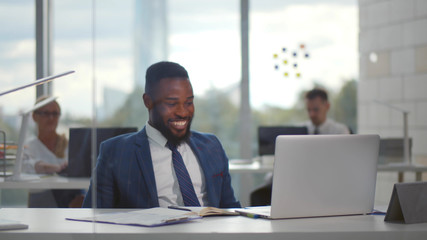 Young african businessman smiling celebrating success while looking at laptop screen in office