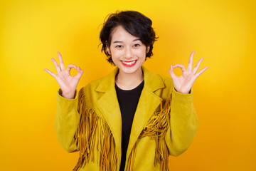 Glad attractive woman shows ok sign with both hands as expresses approval, has cheerful expression. Photo of beautiful female has appealing appearance, being optimistic. Standing against gray wall.