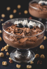 Tasty chocolate mousse dessert placed on dark background with hazelnuts, dark chocolate, blueberries and whipped cream decoration on top. Close up view.
