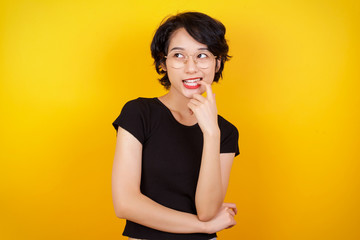 Dreamy female with thoughtful expression, looks to the camera, keeps hand near face, bitting a finger thinks about something pleasant, poses against gray wall.