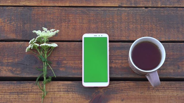 Closeup top view 4k video footage of white smartphone with empty blank green screen laying on wooden brown vintage background outdoors. Cute bouquet of meadow flowers laying near gadget, cup with tea.