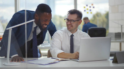 Caucasian and afro businessmen laughing chatting near desktop with laptop