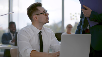 Businessman standing near colleague working on laptop and chatting discussing project