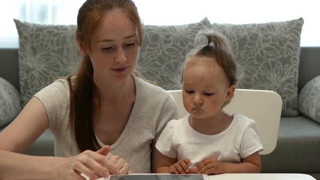 Cute little girl and attractive young mother watch tablet PC. Daughter and mother spend time together and watch images or photos in tablet PC.