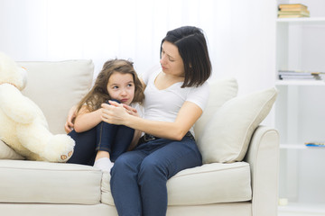Photo of sad little girl sitting on the sofa and her mom hugging her.