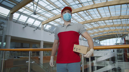 Fototapeta premium Young courier in face mask, gloves and uniform holding parcel standing in empty lobby