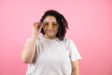Young beautiful woman wearing sunglasses over isolated pink background smiling and taking and lowering her sunglasses