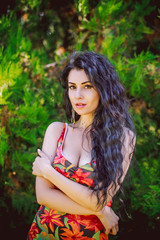 Fototapeta premium A beautiful curly-haired brunette with beautiful breasts in a colorful sundress stands against a background of greenery
