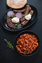 Grilled serbian pljeskavica and cevapi sausages with prebranac and pita, top view on a black stone background, vertical shot
