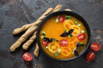 Golden gazpacho soup served in a black bowl with salted breadsticks, elevated view on a brown stone background, studio shot