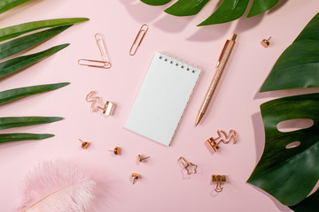 Flat lay of blank notepad, golden office supplies and palm leaves on pink background