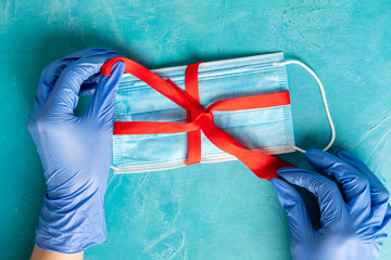 Stay safe. Present surprise. Hands in medicine gloves taping face mask with red ribbon. Textured blue background. COVID-19 prevention. Quarantine measures. Epidemic self-isolation
