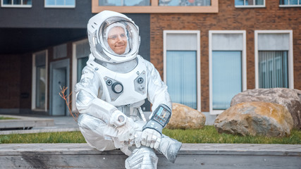 Happy Man in Spacesuit is Sitting on Wooden Bench. Posittive Astronaut Looks in the Distance. Emotionally Joyful Spaceman in White Futuristic Suit with Technological Panel on His Hand. © Gorodenkoff